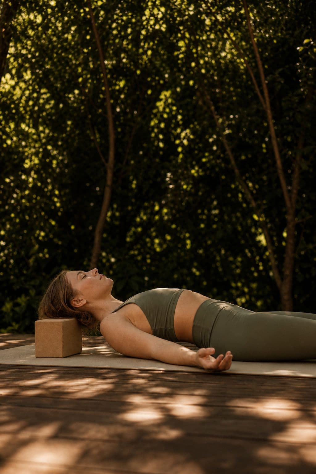 A woman resting in savasana on an outdoor deck in dappled light